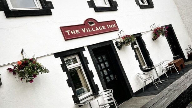 Un bâtiment blanc avec une enseigne indiquant « The Village Inn ». On y voit des jardinières suspendues et un banc en bois à l’extérieur.