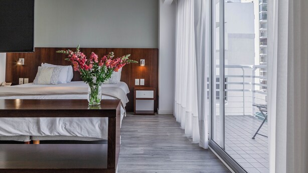 A hotel room with a bed, bedside tables, a nightstand, a vase with flowers, and a view of the outside balcony. - Color Botanico (Buenos Aires)