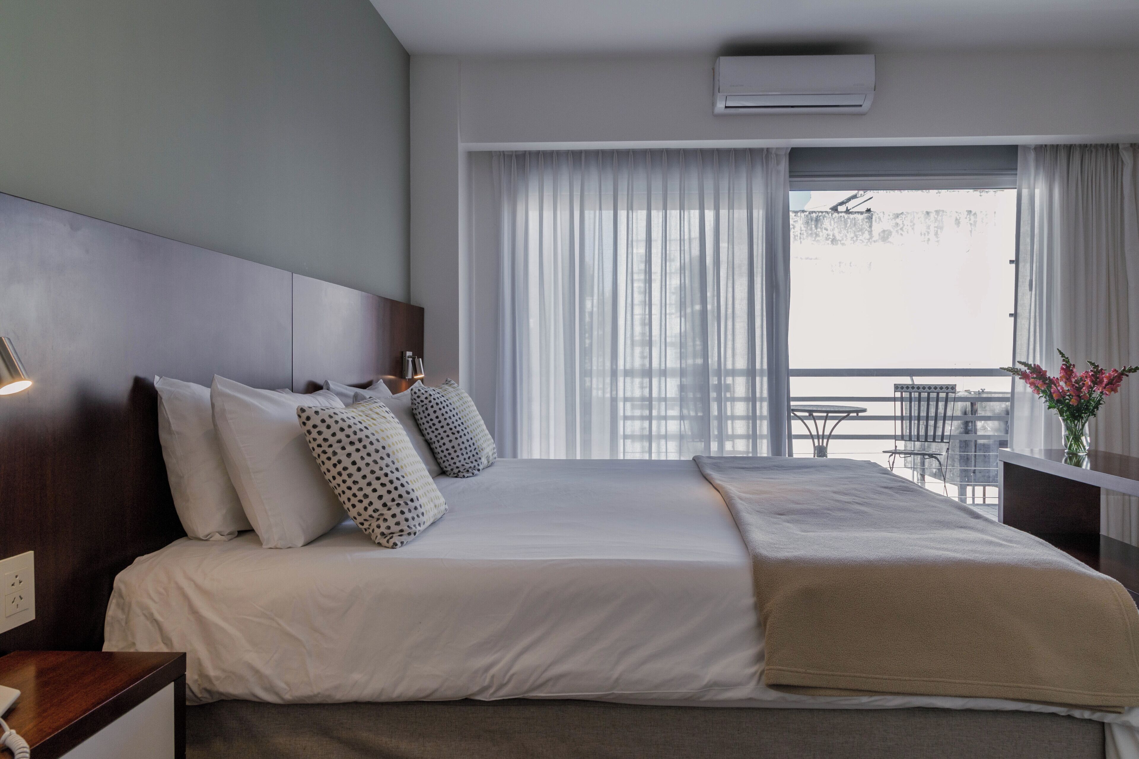A hotel room with a bed, a bedside table with a phone, a window with curtains, and a balcony with chairs.