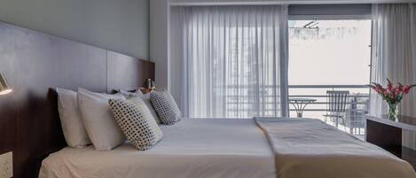 A hotel room with a bed, a bedside table with a phone, a window with curtains, and a balcony with chairs.