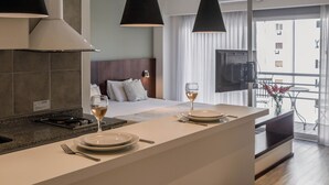 A modern hotel room with a kitchen area, a dining table with plates and glasses, a bed with a headboard, and a TV mounted on the wall. - Color Botanico (Buenos Aires)