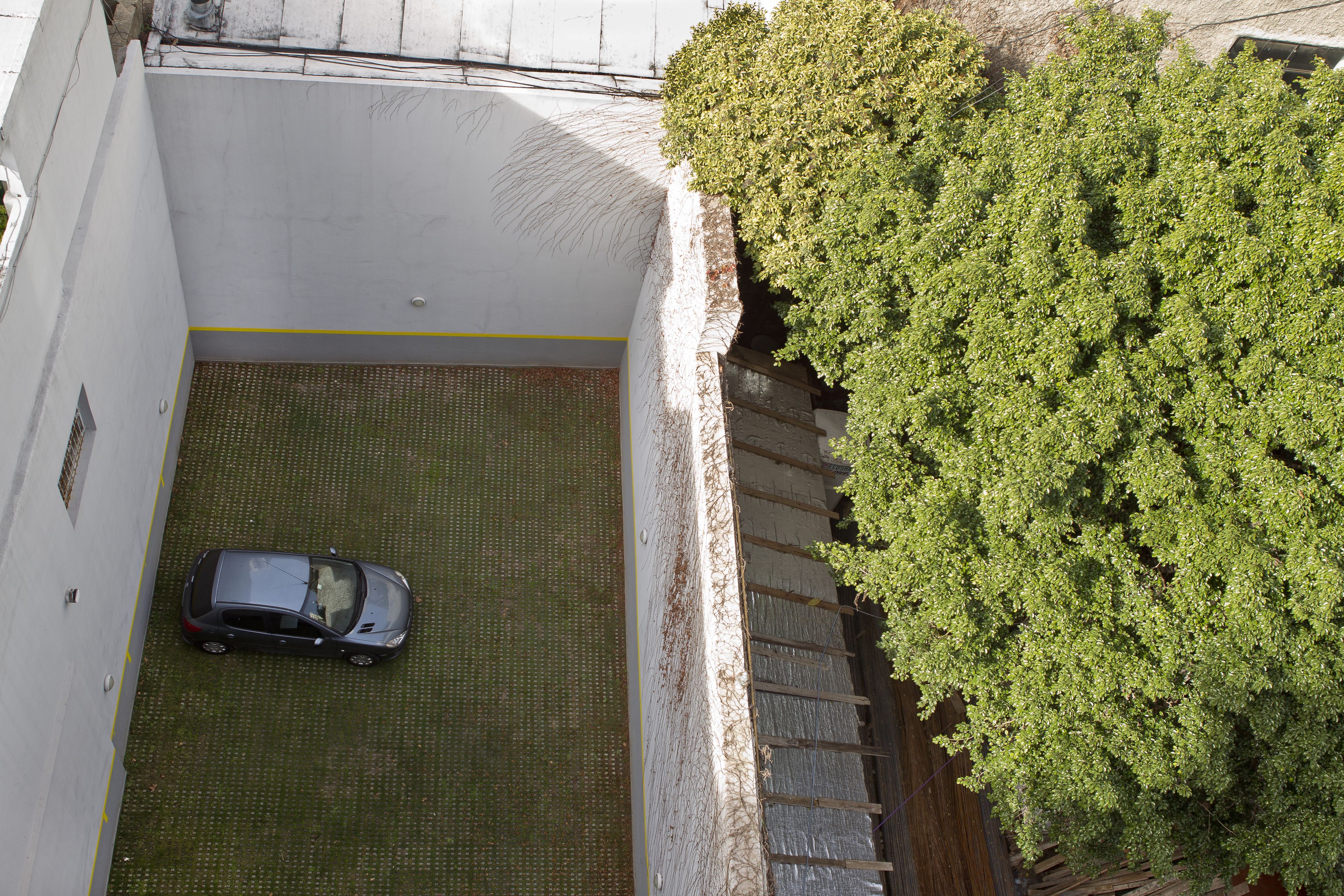 A car parked in a covered parking area with a grassy surface and a tree to the right.