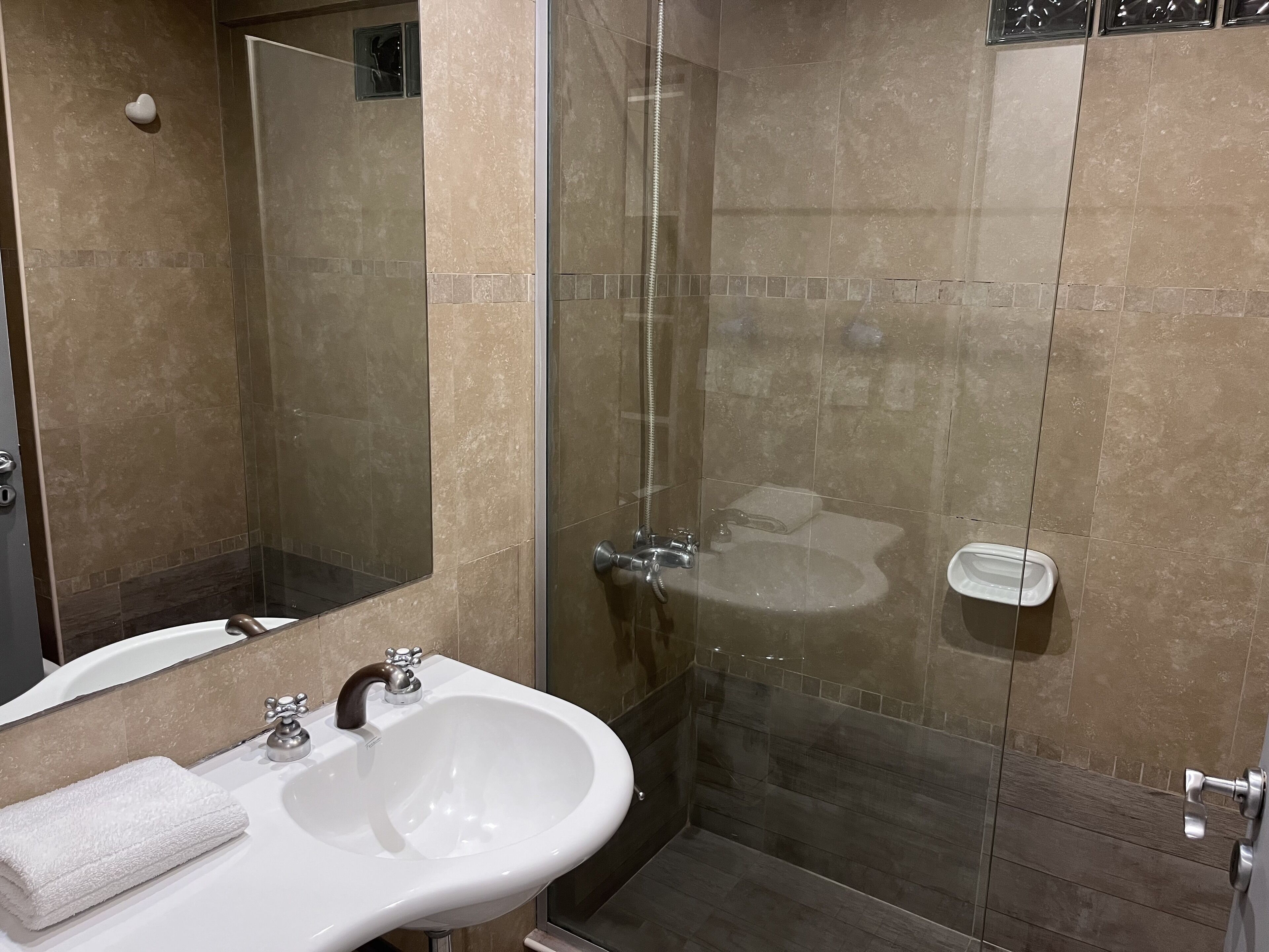A modern bathroom with a glass-enclosed shower, a white sink with a mirror, and a small bedside table with a towel.