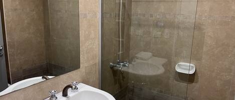 A modern bathroom with a glass-enclosed shower, a white sink with a mirror, and a small bedside table with a towel.
