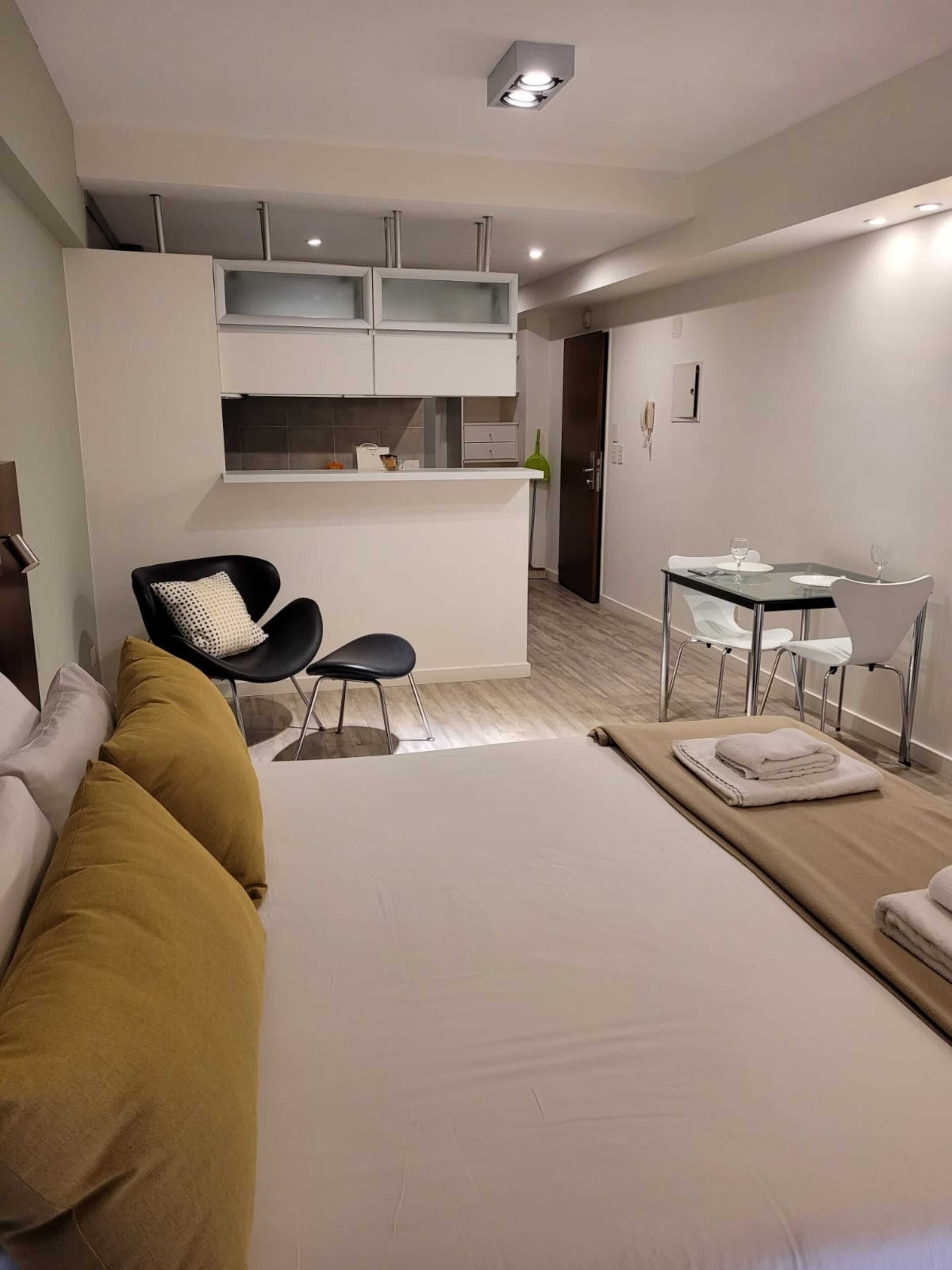 Una camera d'hotel moderna con un letto, una poltrona, un tavolo da pranzo con sedie, un angolo cottura e una porta che dà su un'altra stanza.