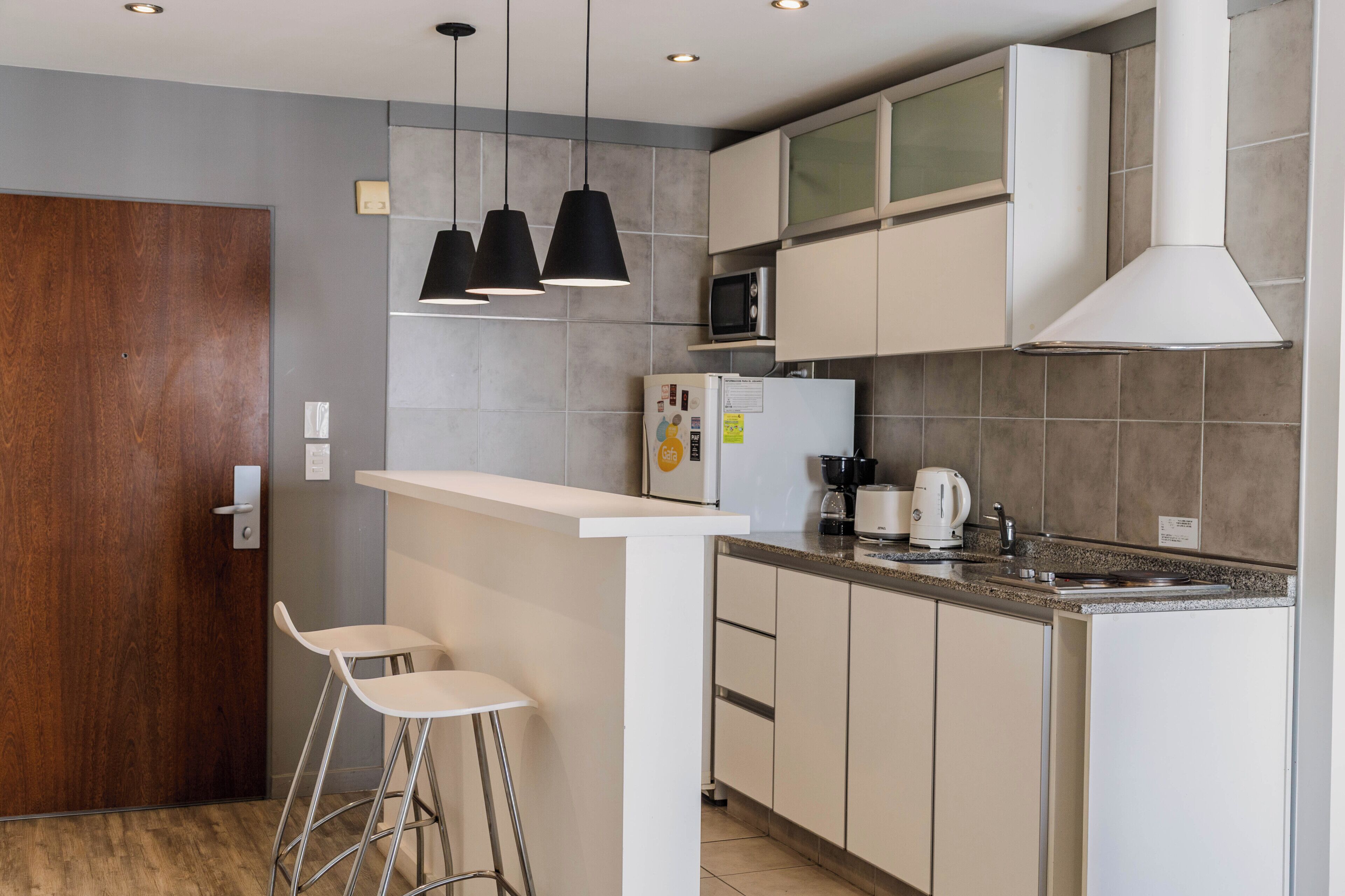 Una cocina moderna con encimera blanca, taburetes de bar, microondas, cafetera y luces colgantes.