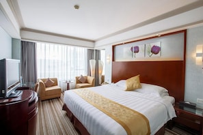 Ein Hotelzimmer mit einem großen Bett, zwei Sesseln, einem Schreibtisch mit Fernseher und einem großen Fenster mit Vorhängen.