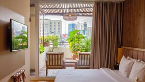Habitación de hotel con cama, televisor montado en la pared, balcón con sillas y plantas, y vista a la ciudad.