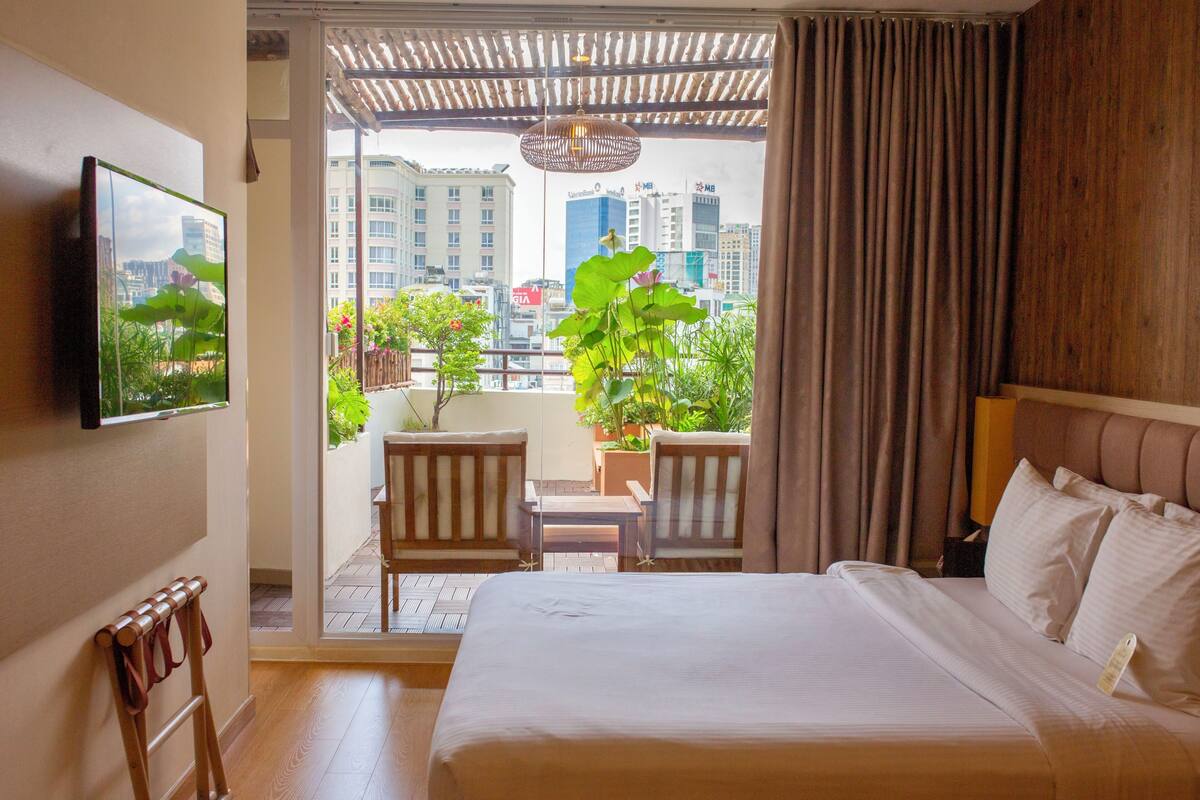 A hotel room with a bed, a TV mounted on the wall, a balcony with chairs and plants, and a city view.