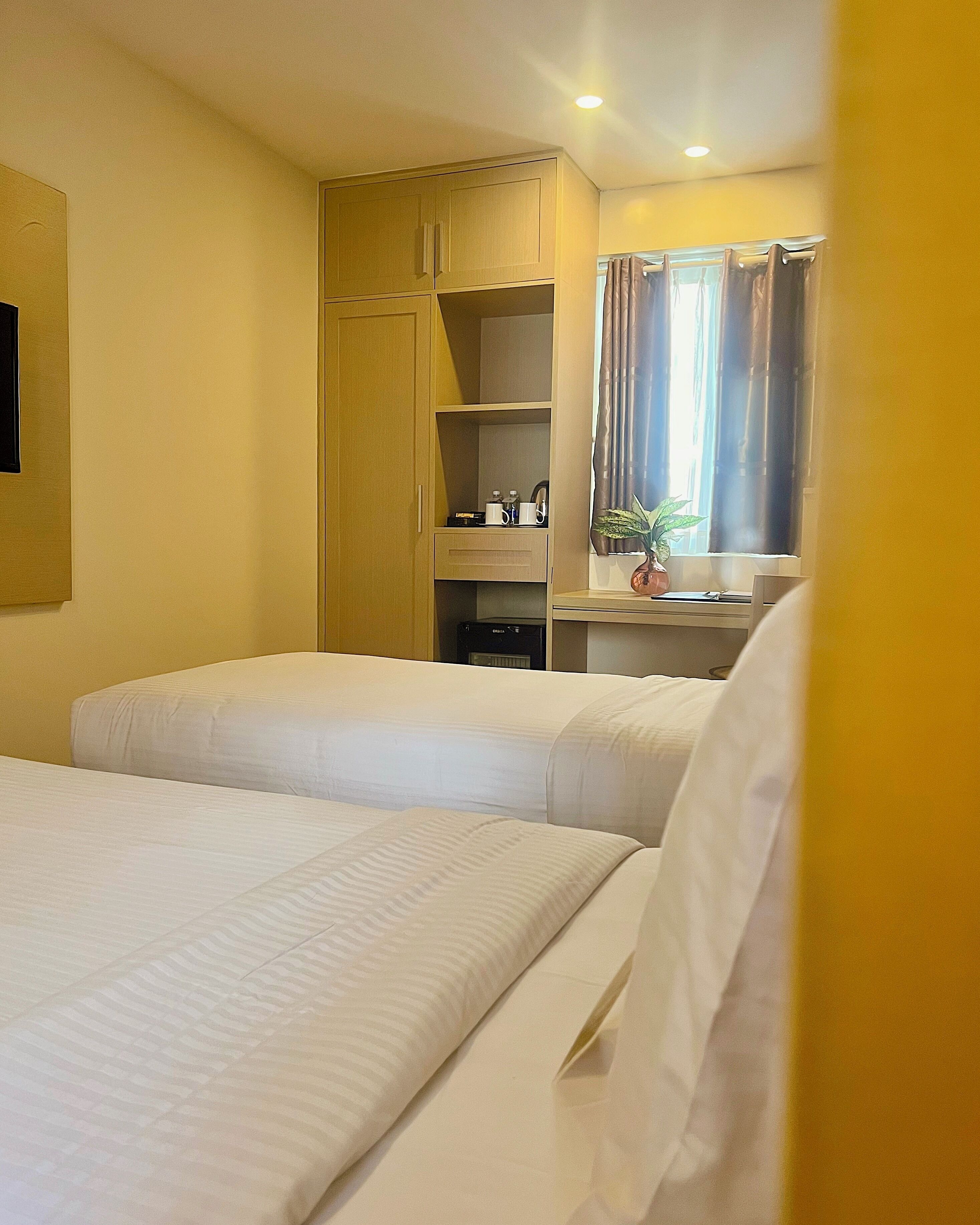 A hotel room with two beds, a wardrobe, a desk with a plant, and a window with curtains.