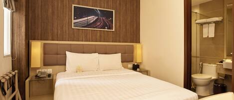 A hotel room with a large bed, a wooden panel wall, a framed picture, a bedside table with a lamp, and a bathroom visible through an open door.