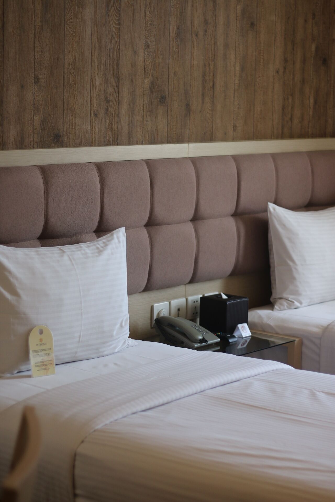 A hotel room with a wooden headboard, two beds with white bedding, a bedside table with a telephone and a black object, and a wall-mounted electrical outlet.