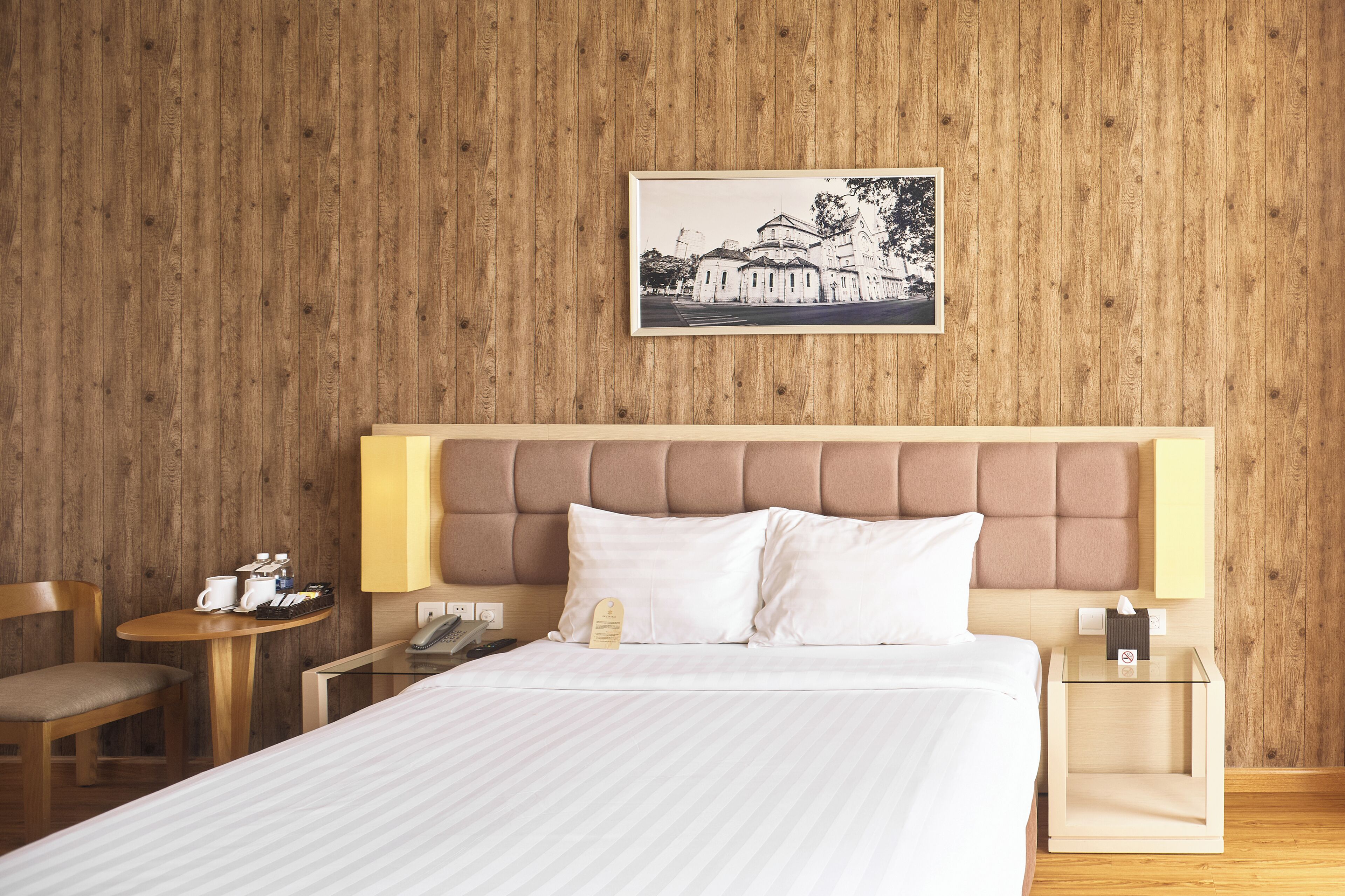 Habitación de hotel con pared de madera, una cama con ropa blanca, cabecera, mesitas de noche, una silla y un cuadro en la pared.