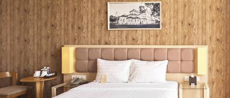 Habitación de hotel con pared de madera, una cama con ropa blanca, cabecera, mesitas de noche, una silla y un cuadro en la pared.