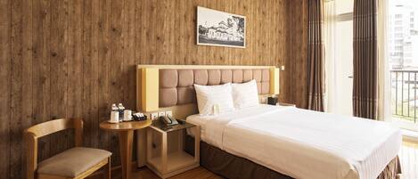 A hotel room with a wooden wall, a bed with white bedding, a wooden chair, a bedside table with a lamp, and a window with curtains.