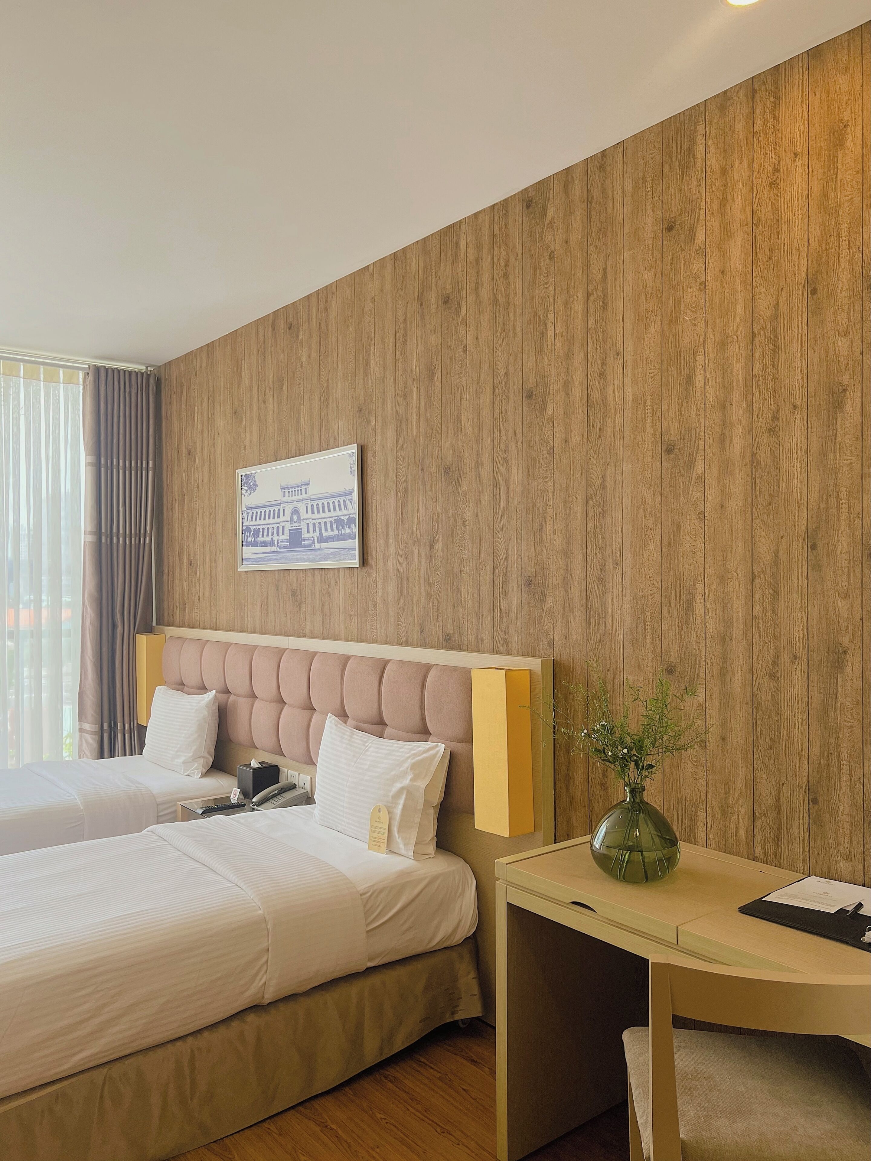 A hotel room with two beds, a wooden wall, a framed picture, a bedside table with a vase, and a window with curtains.