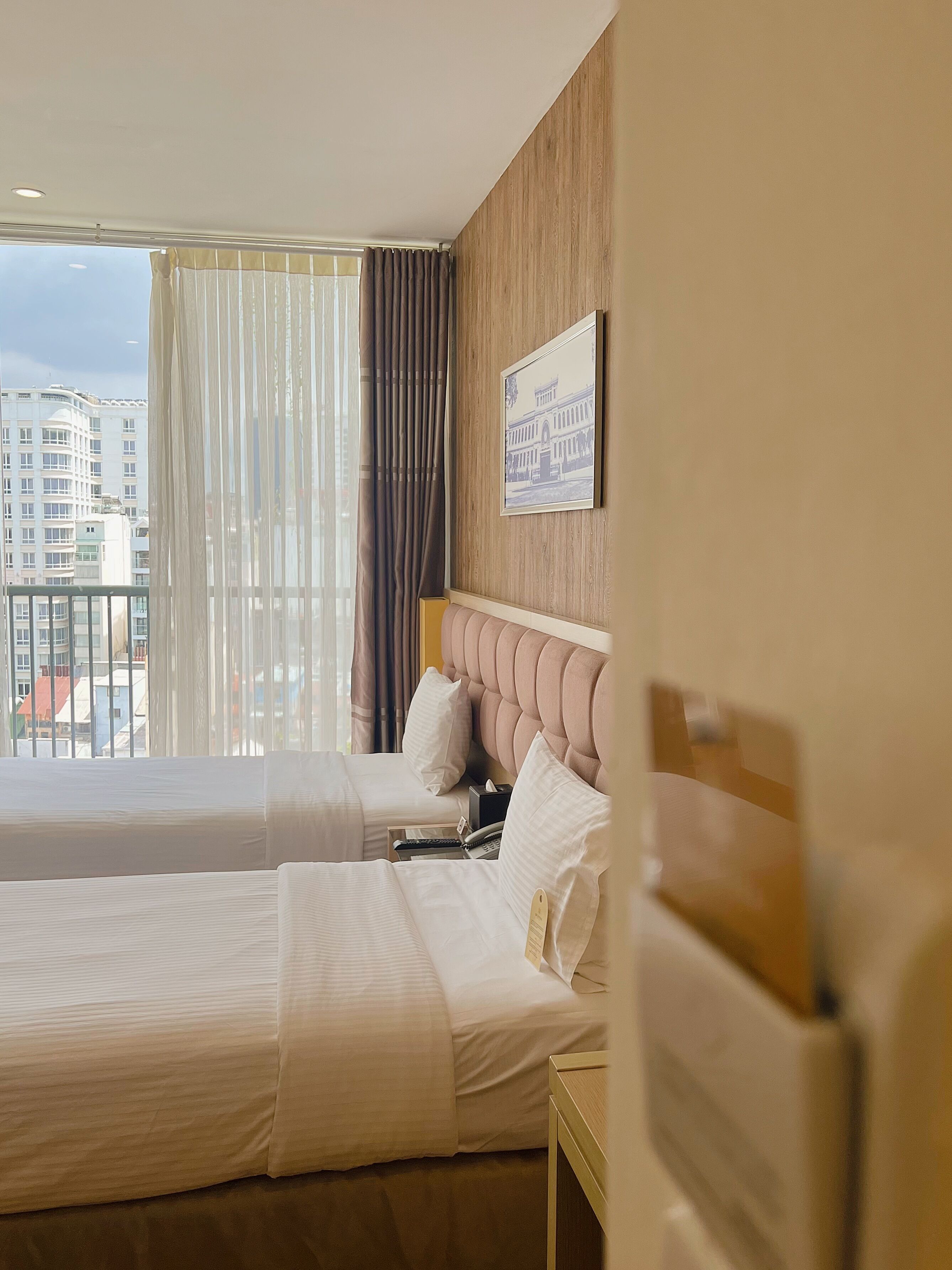 A hotel room with two beds, a wooden headboard, a window with curtains, and a city view.