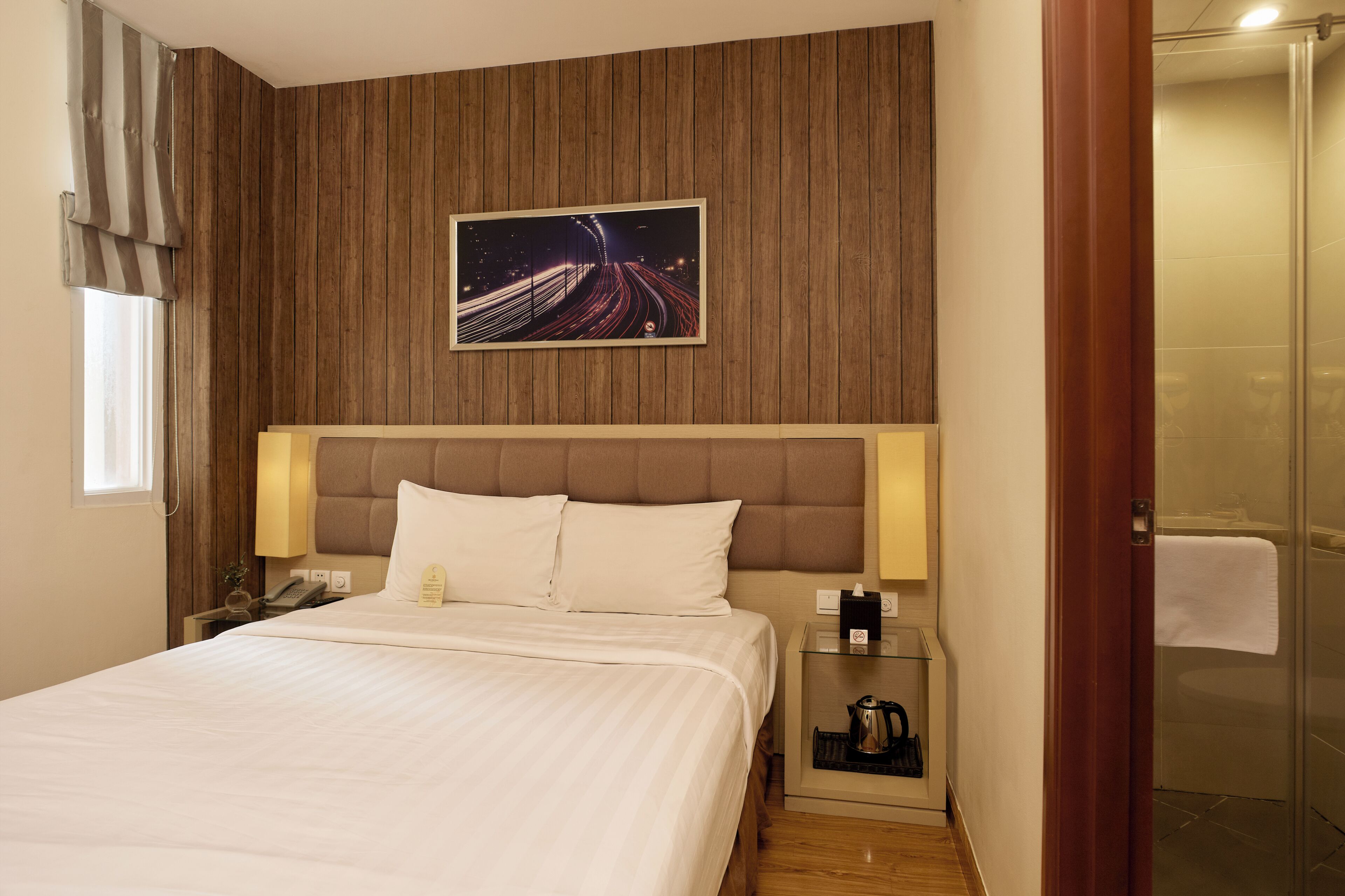 A hotel room with a bed, a nightstand, a telephone, a lamp, a picture of a bridge, and a shower area.