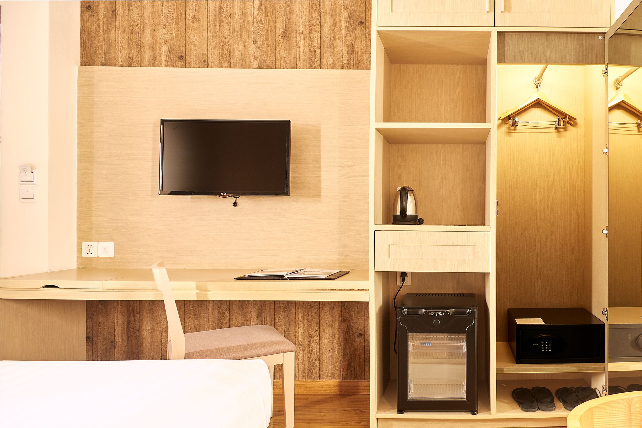 A hotel room with a flat-screen TV mounted on the wall, a desk with a chair, a coffee maker, a microwave, and a shoe rack.