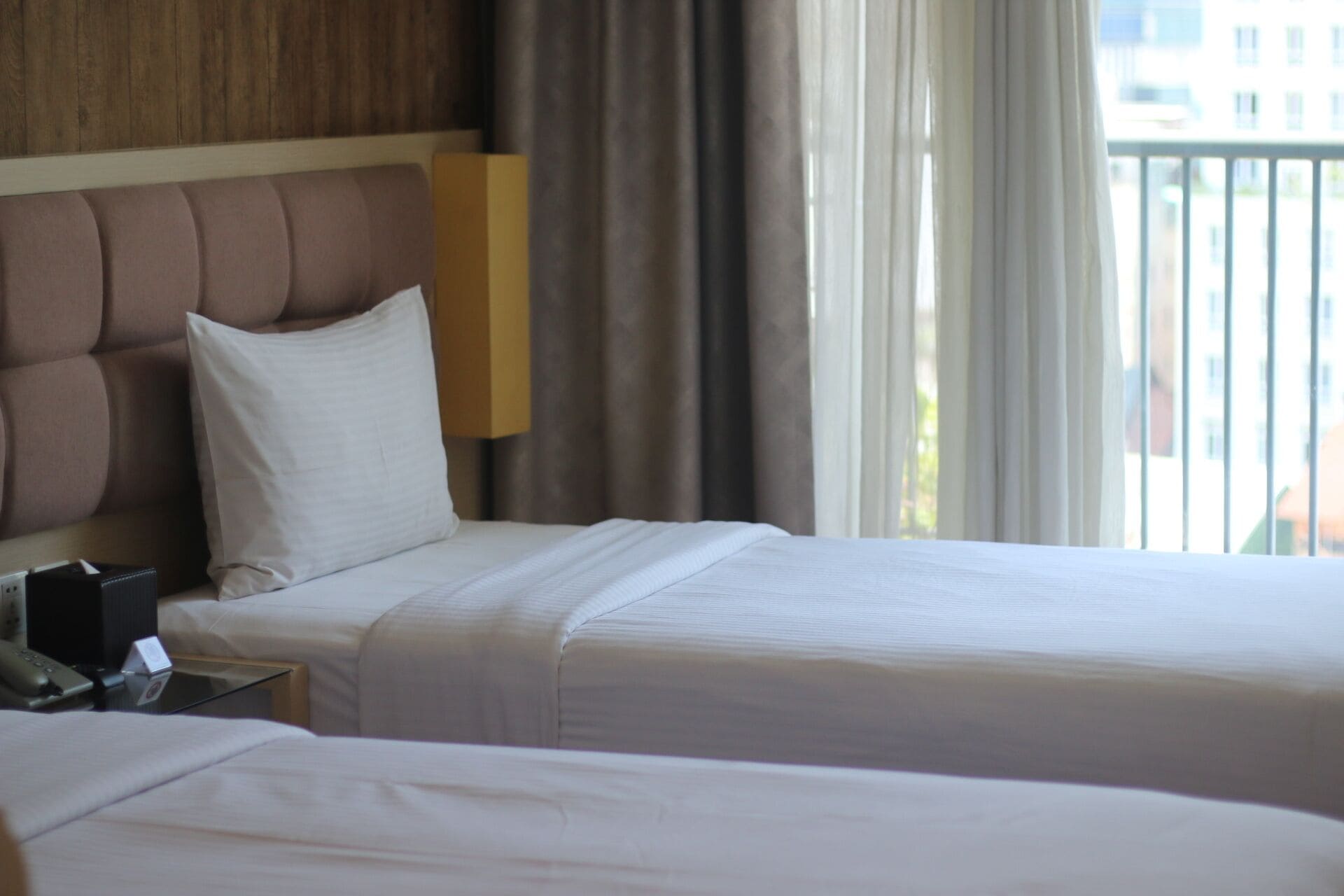 Habitación de hotel con dos camas, cabecero de madera, mesita de noche con teléfono y vista al exterior a través de una ventana con cortinas.