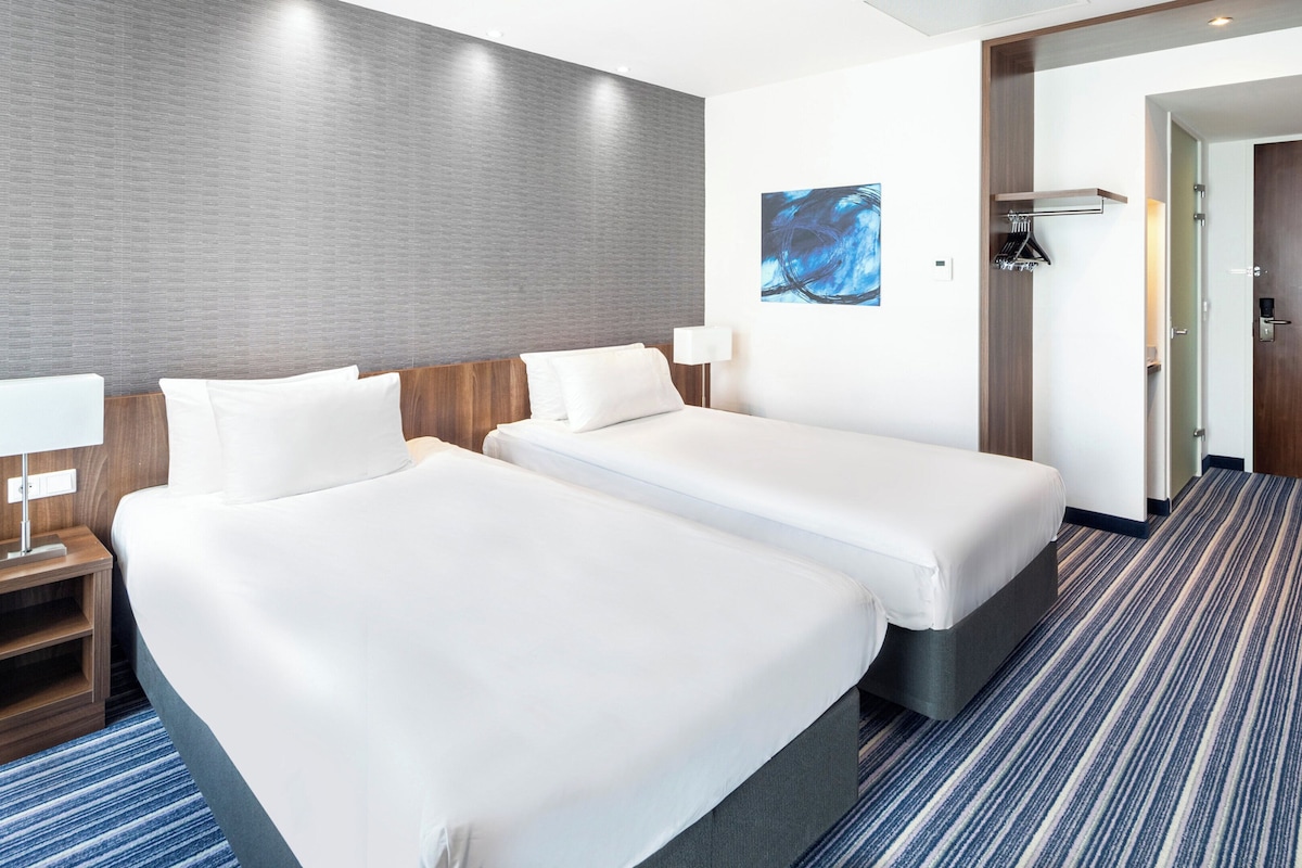 A hotel room with two beds, a nightstand with a lamp, a coat rack, and a door leading to another room.
