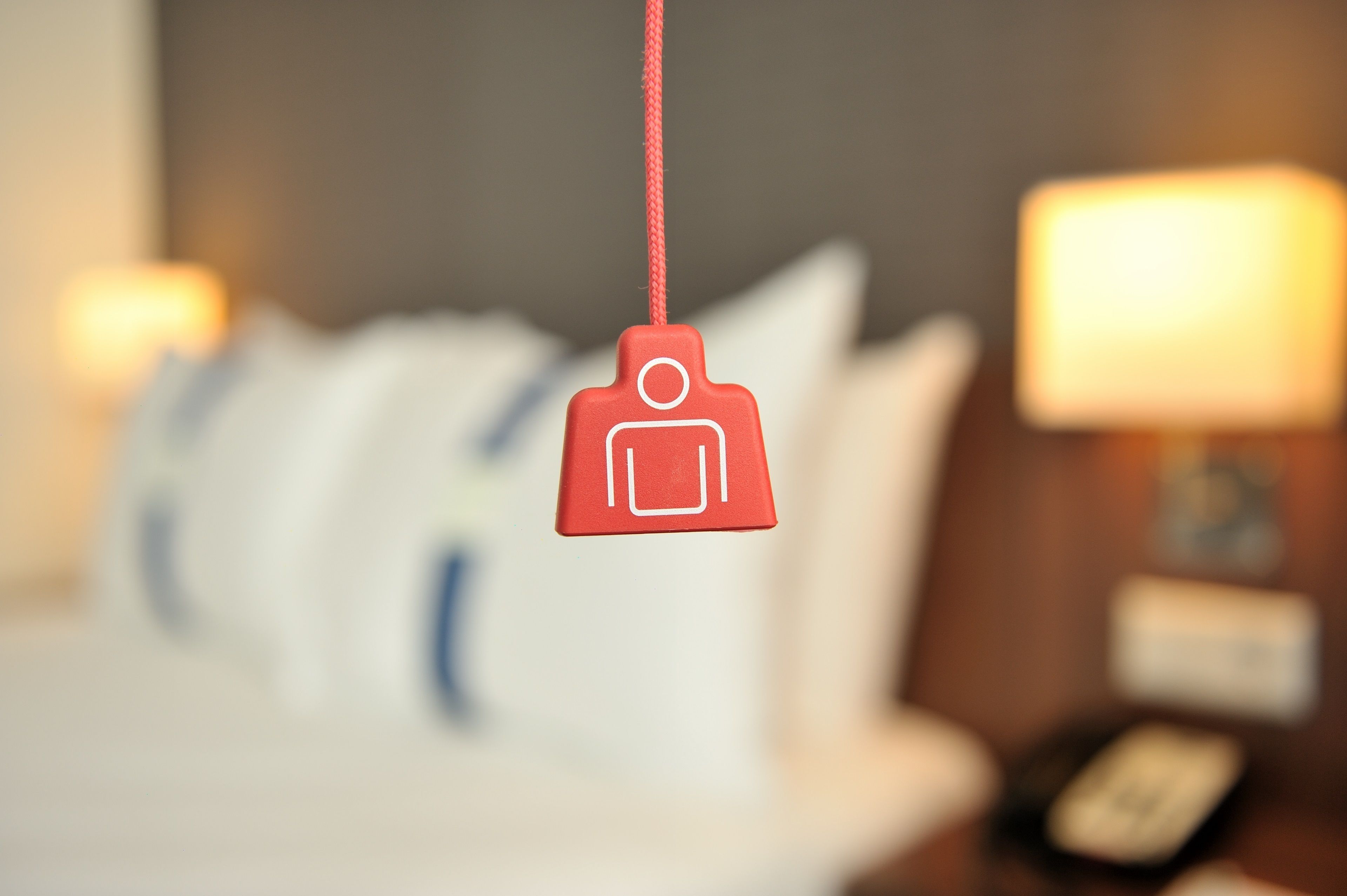 A blurred hotel room with a hanging tag and a lamp on a bedside table.