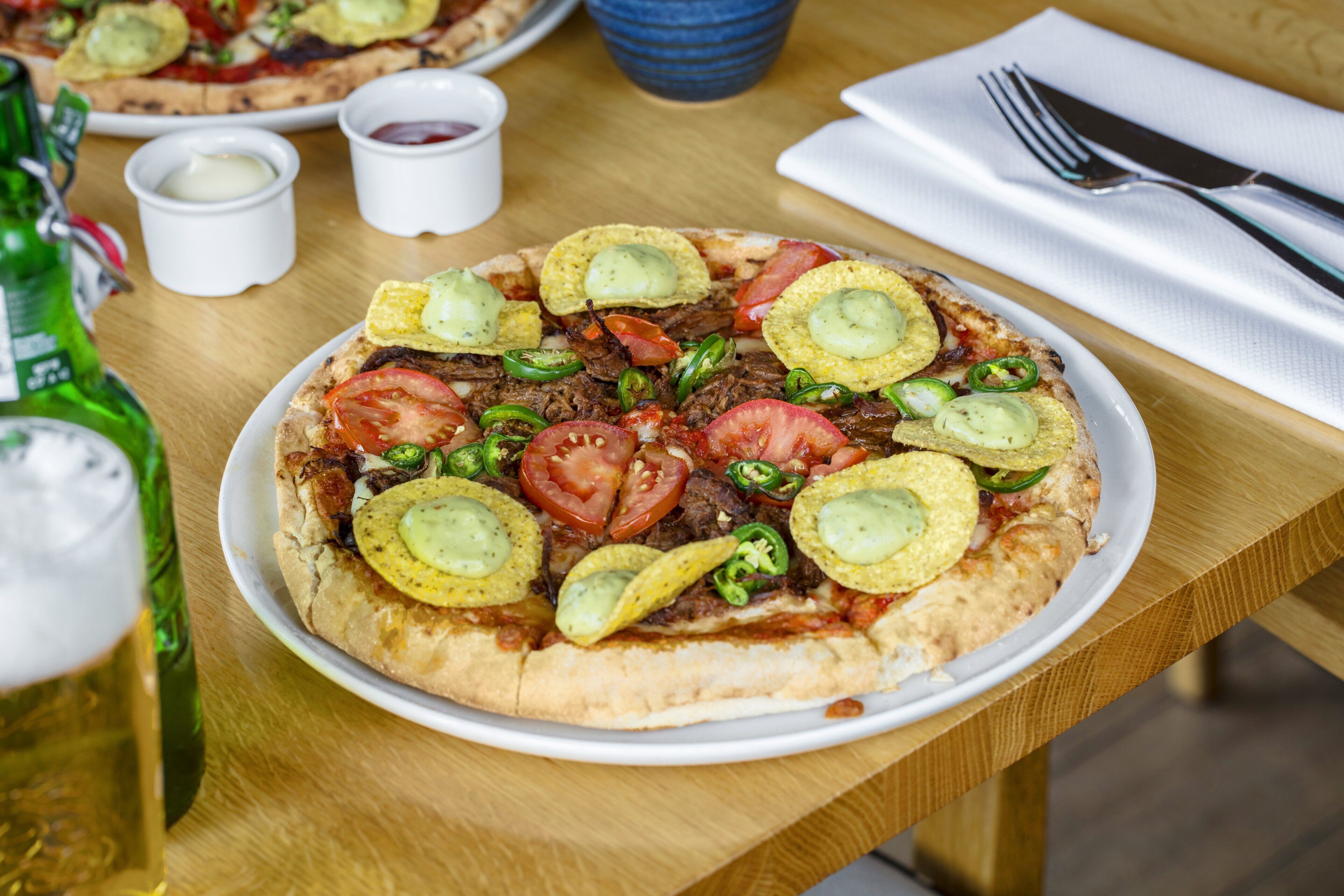 Pizza met verschillende toppings op een wit bord, vergezeld van kleine schaaltjes met sauzen en een vork op een servet.