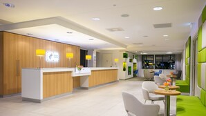 Un espace d’accueil moderne, avec un comptoir en bois, un coin salon vert et un restaurant visible au fond.