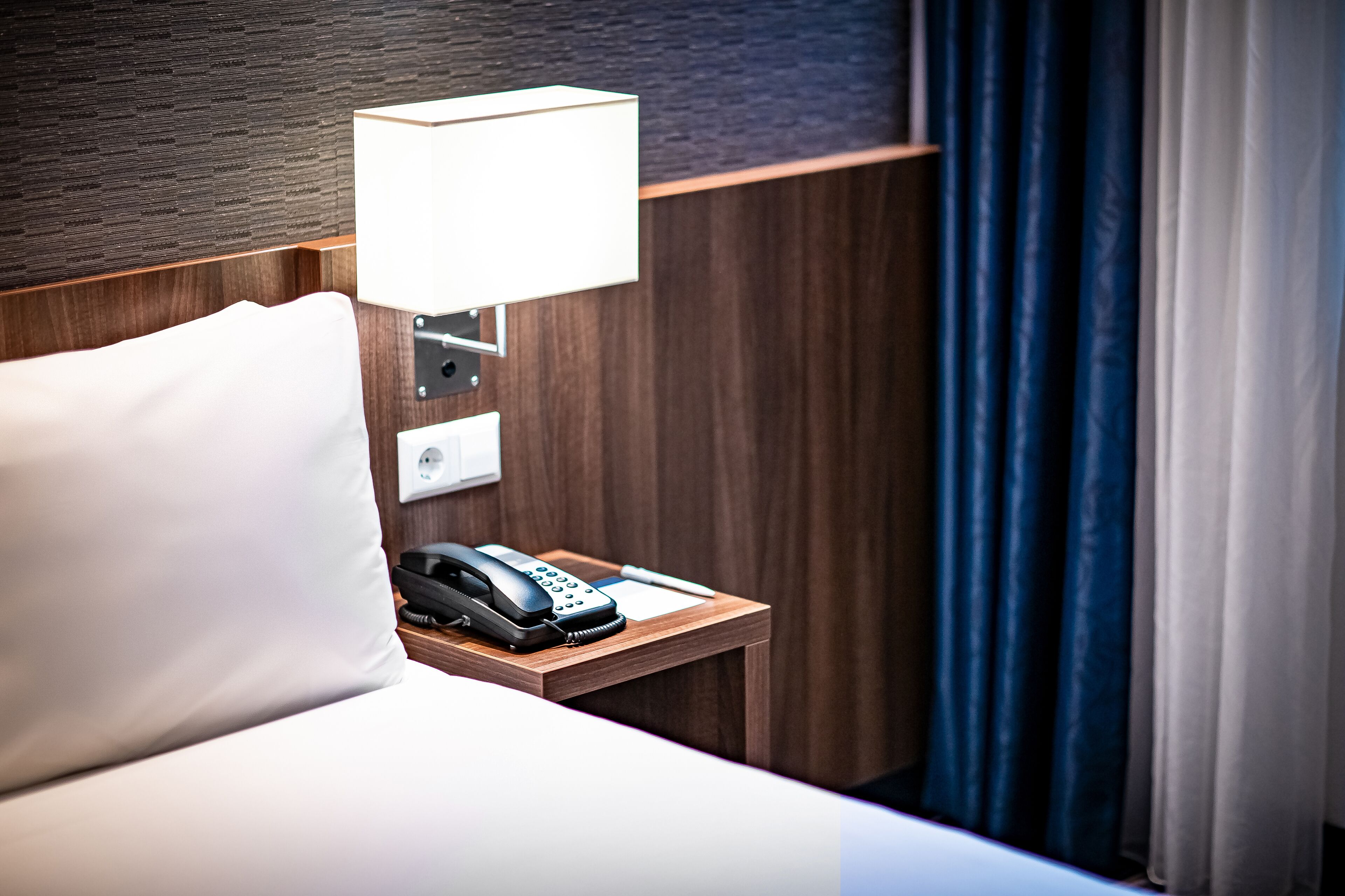 A hotel room with a wooden headboard, a bedside lamp, a telephone, and a white pillow.