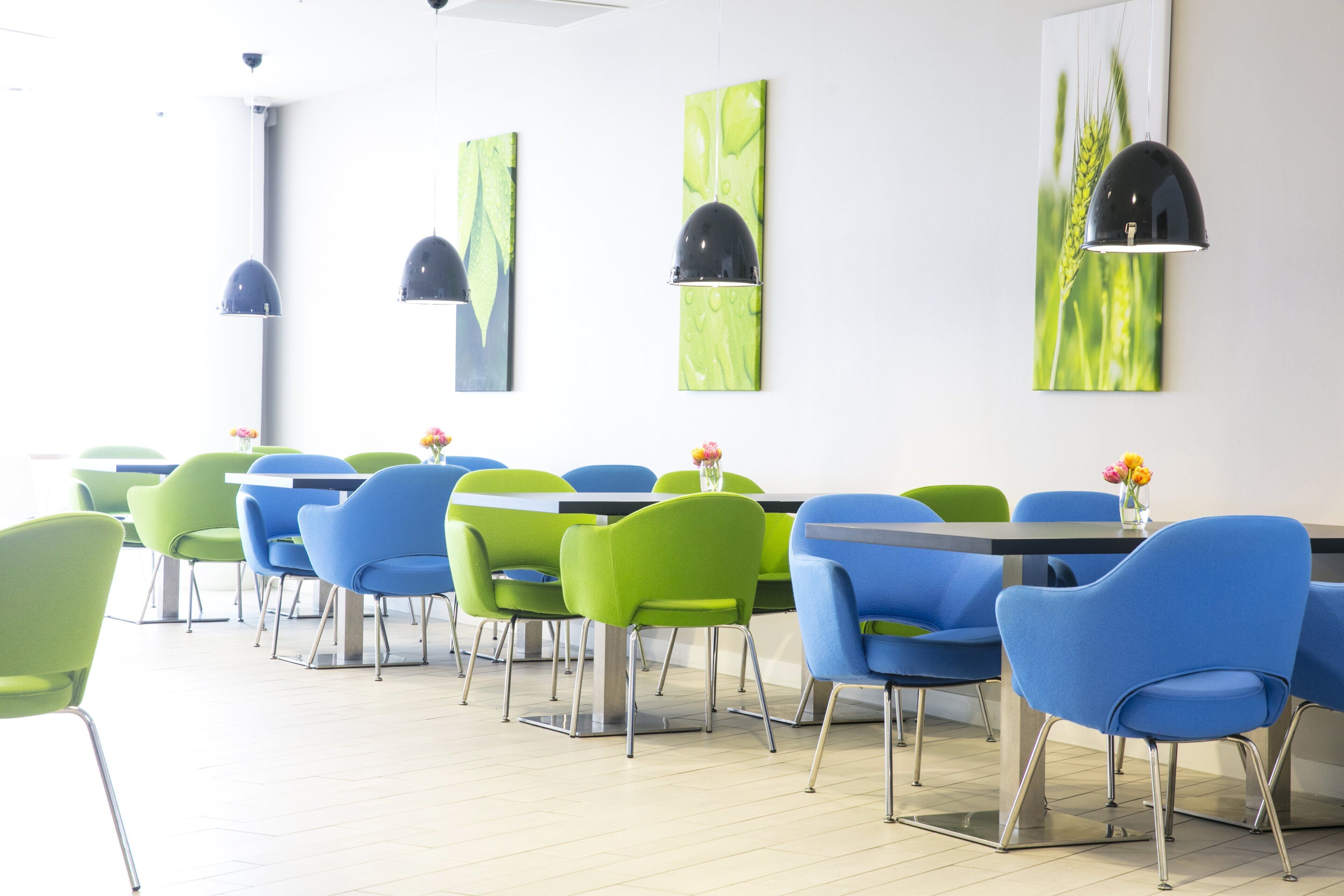 A modern dining area with green and blue chairs, white tables, and abstract green wall art.