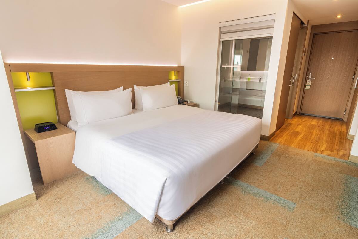 A hotel room with a large bed, bedside tables, a nightstand, a glass door leading to a bathroom, and a wooden floor.