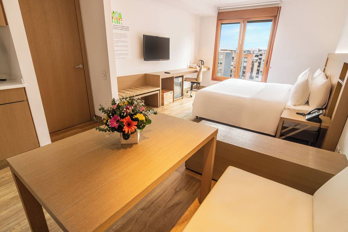 A hotel room with a bed, a desk, a chair, a TV, and a window with a city view.