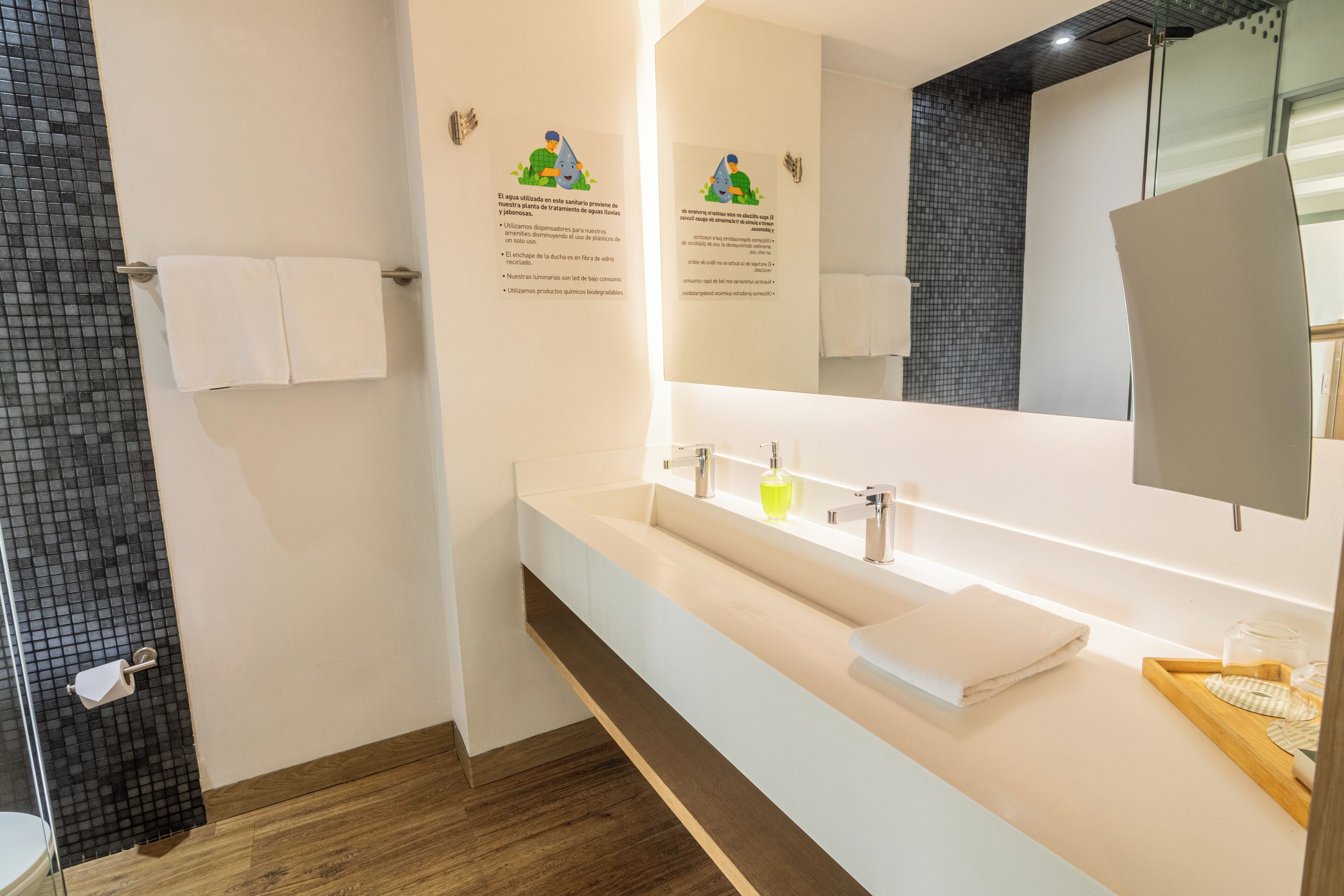 Un baño moderno con un espejo grande, un lavabo blanco y suelo de madera. Hay dos toallas colgadas en la pared y un box de ducha de vidrio.
