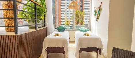 A modern hotel room with two beds, a reception desk, and large windows offering a city view.
