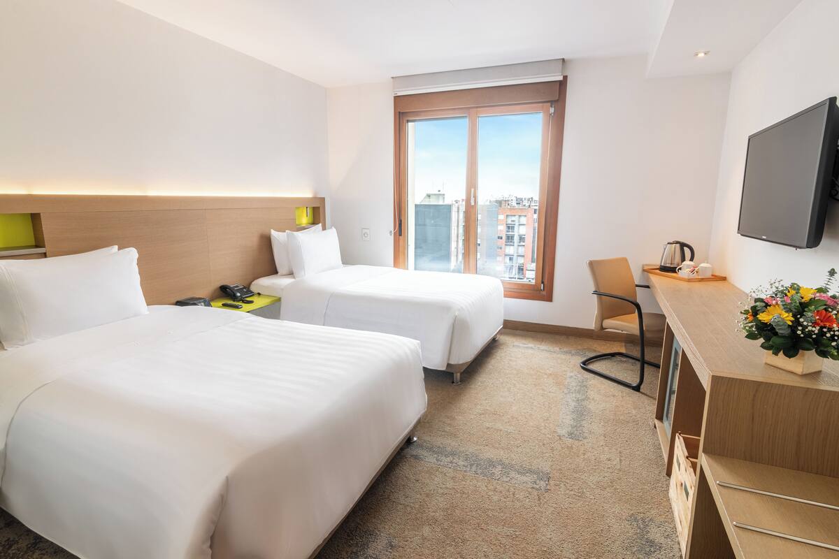 A hotel room with two beds, a desk, a chair, a TV, and a window with a city view.
