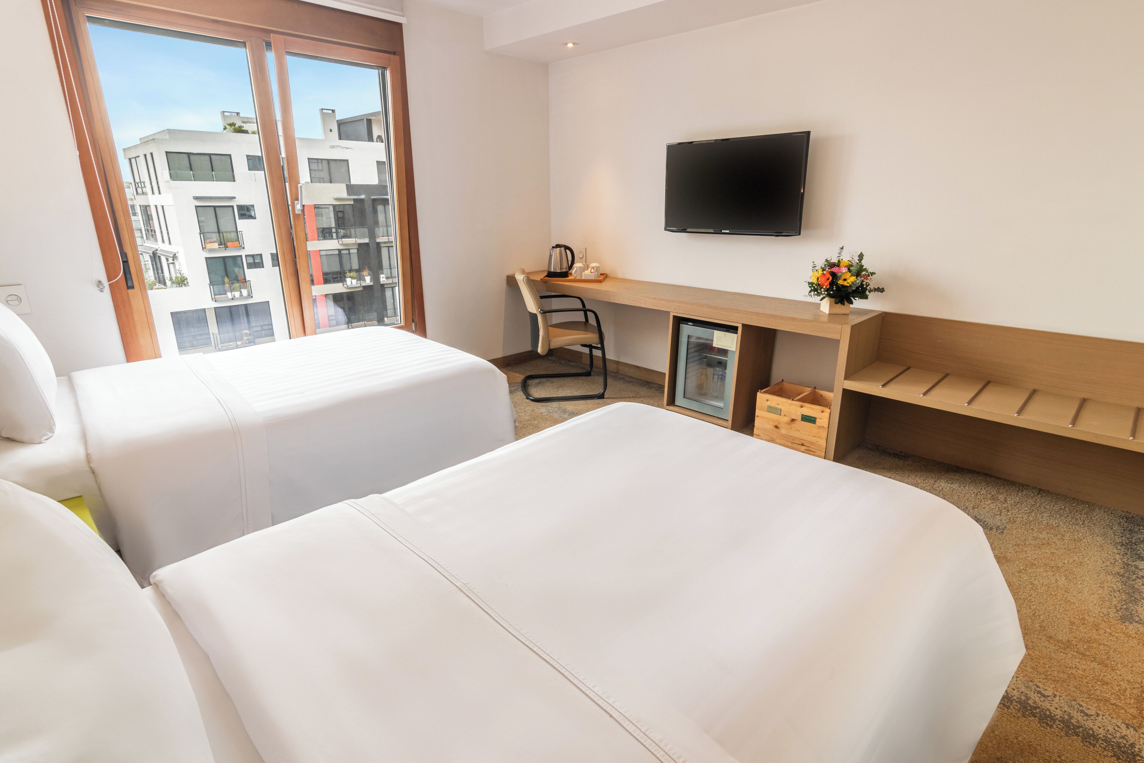 A hotel room with two beds, a desk with a chair, a TV, and a small table with flowers.