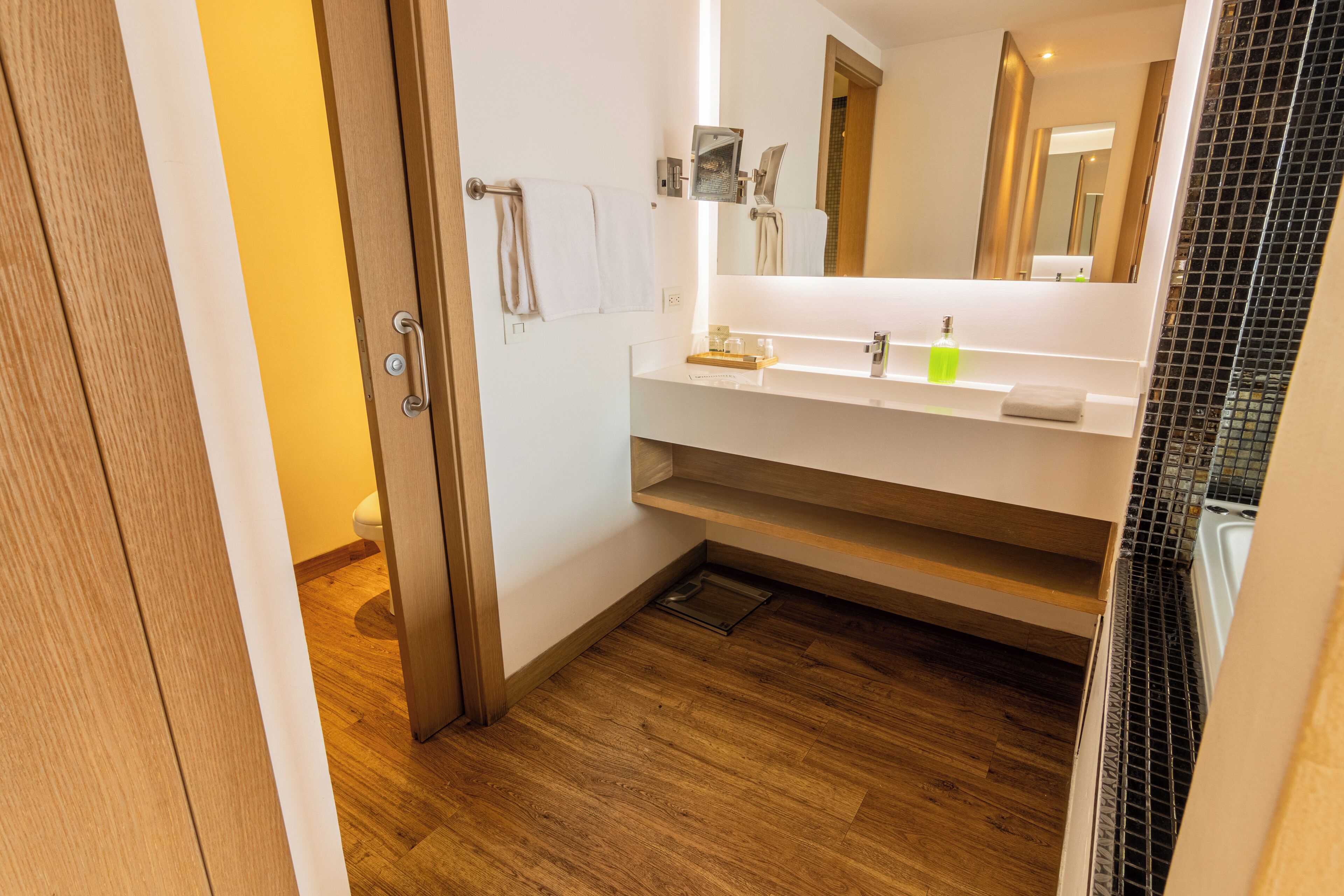 Een moderne badkamer met een houten vloer, witte wanden, een grote spiegel en een witte wastafel met zeepdispenser.