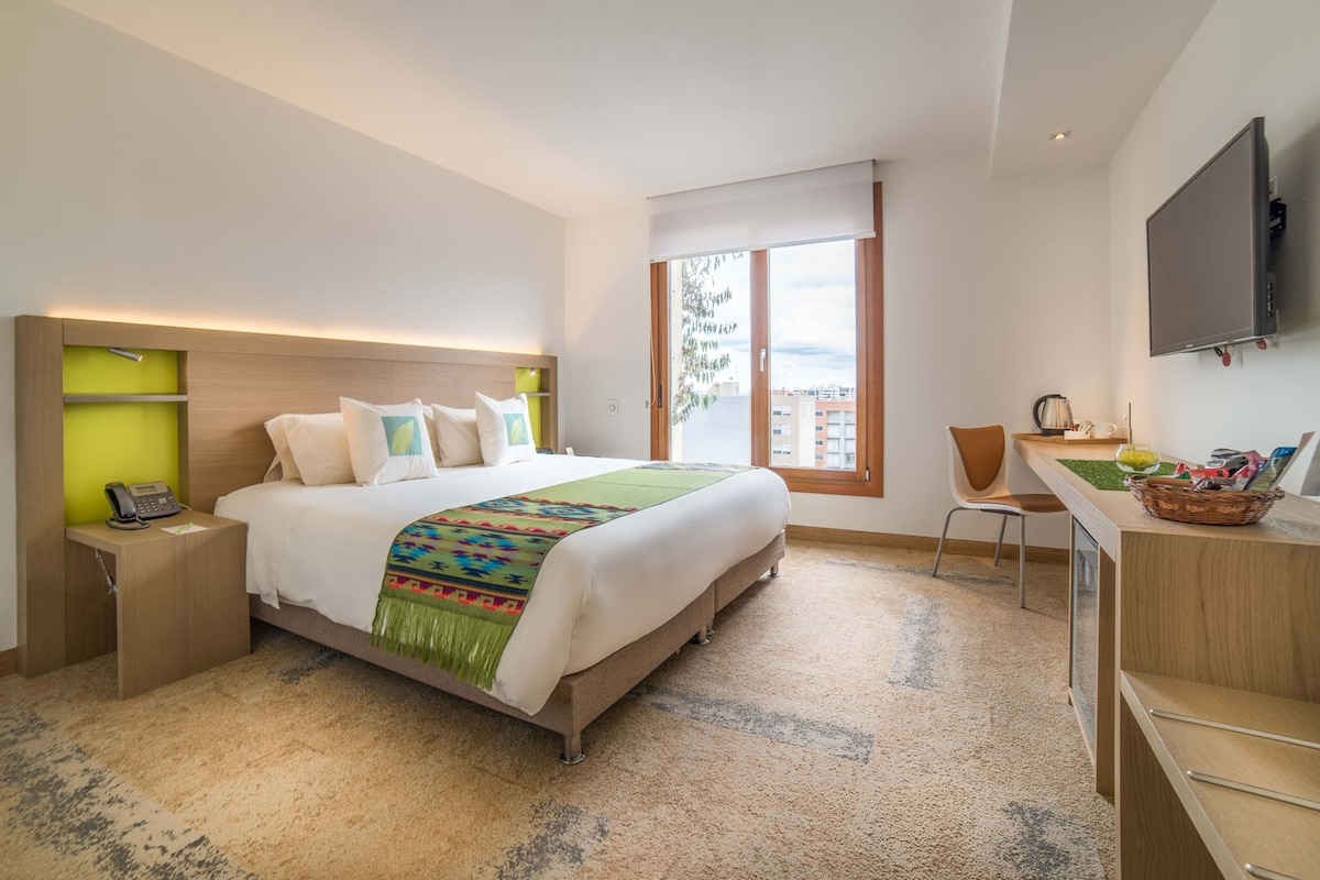 A hotel room with a bed, bedside table, chair, desk, and a TV mounted on the wall.