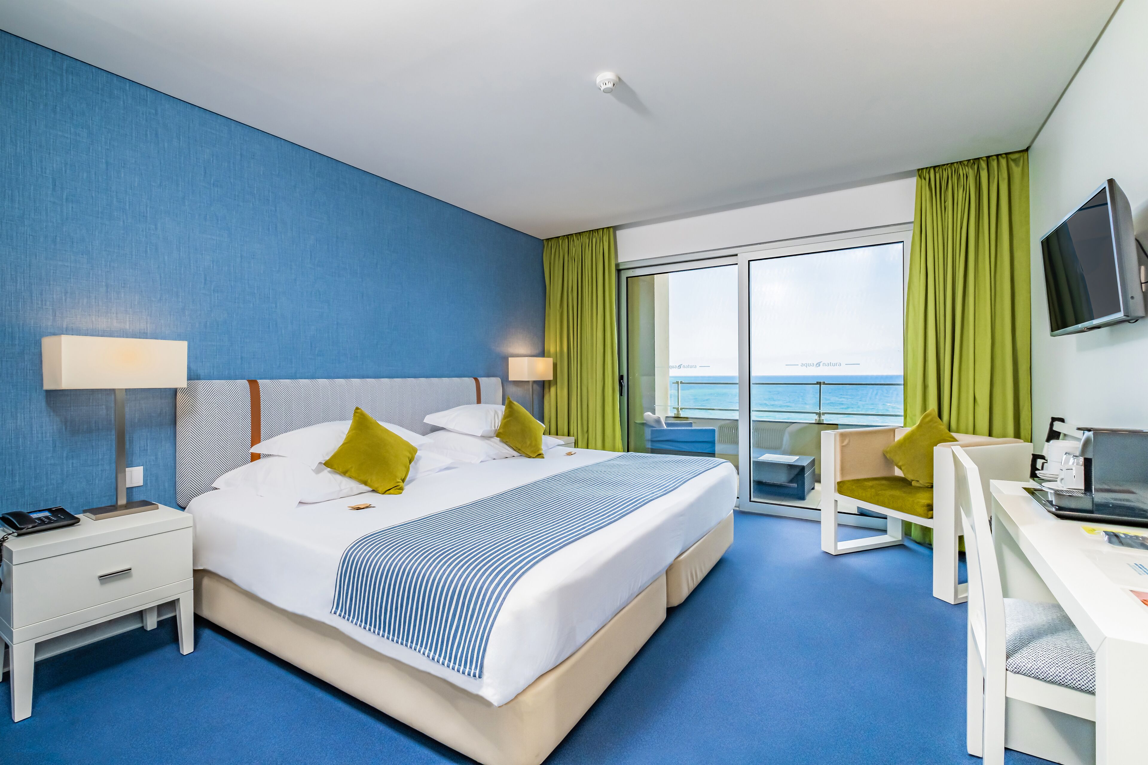 A modern hotel room with a large bed, a desk, and a view of the sea.