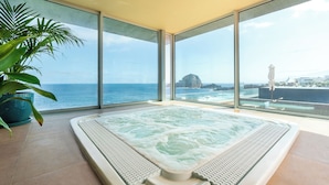 Un jacuzzi grande y rectangular con borde texturizado, situado en una habitación con ventanas amplias que ofrecen vista al océano y una isla distante.