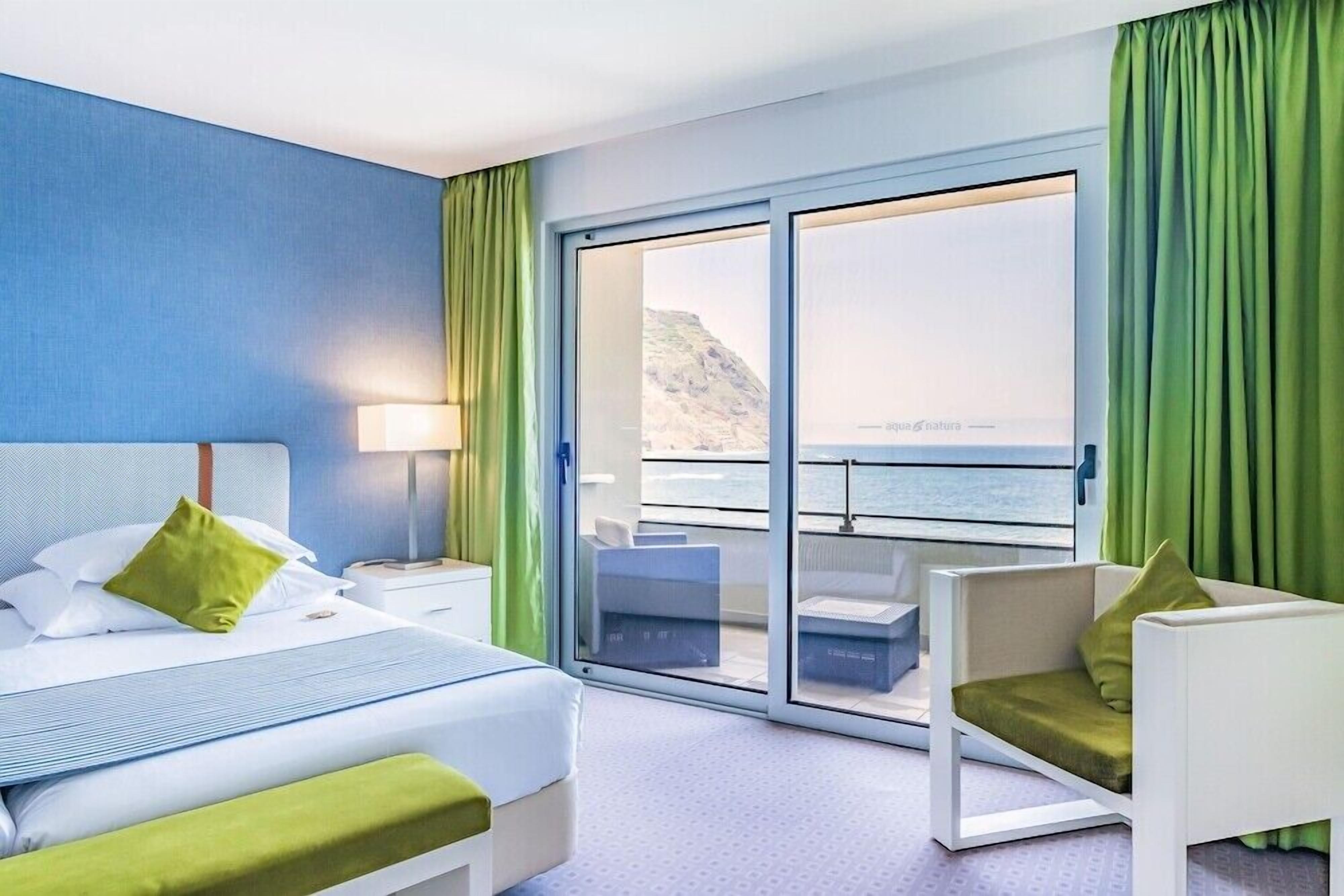 A hotel room with a bed, a chair, a nightstand, a lamp, a sliding glass door leading to a balcony with a view of the sea.