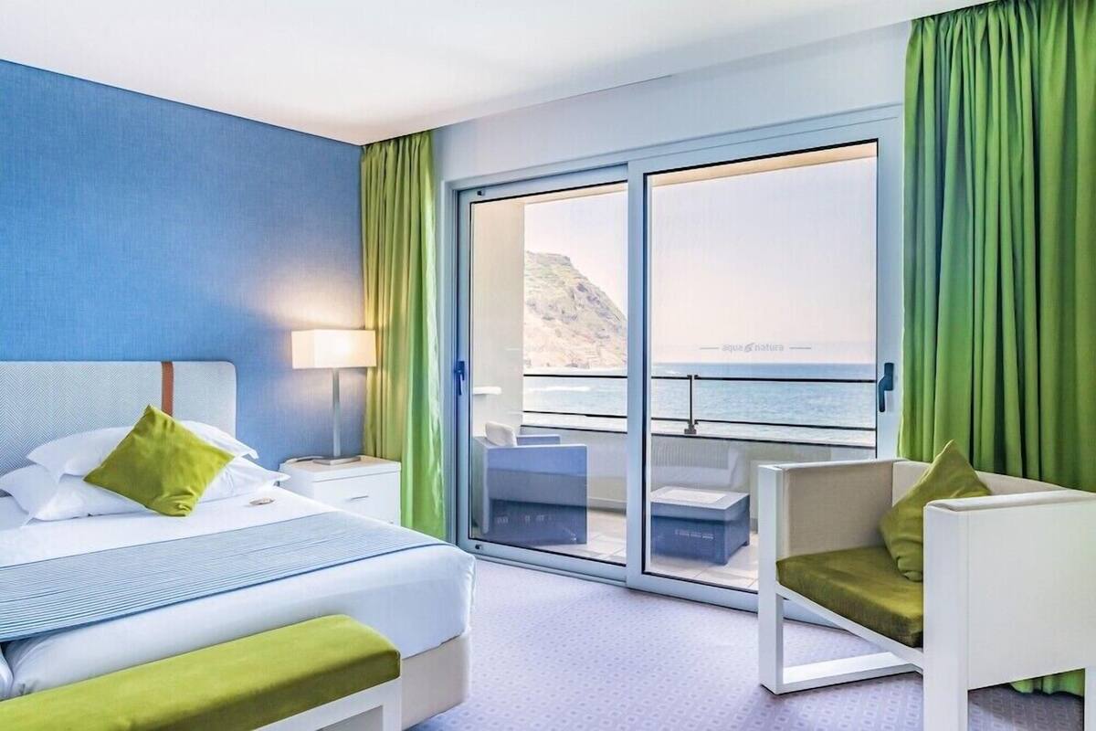 A hotel room with a bed, a chair, a nightstand, a lamp, a sliding glass door leading to a balcony with a view of the sea.