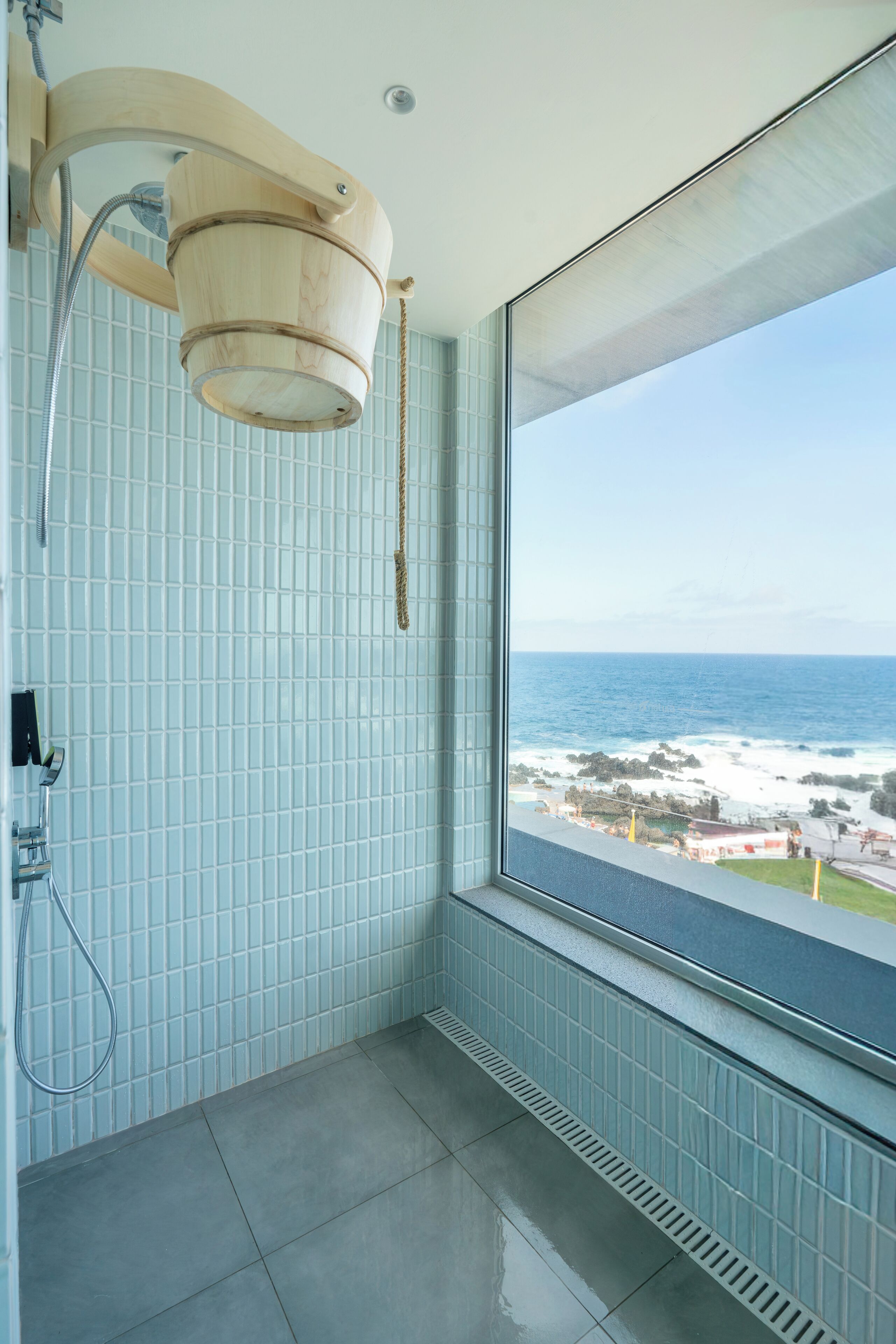 Un baño con vista al océano, un cabezal de ducha en forma de cubo de madera y paredes de azulejos.