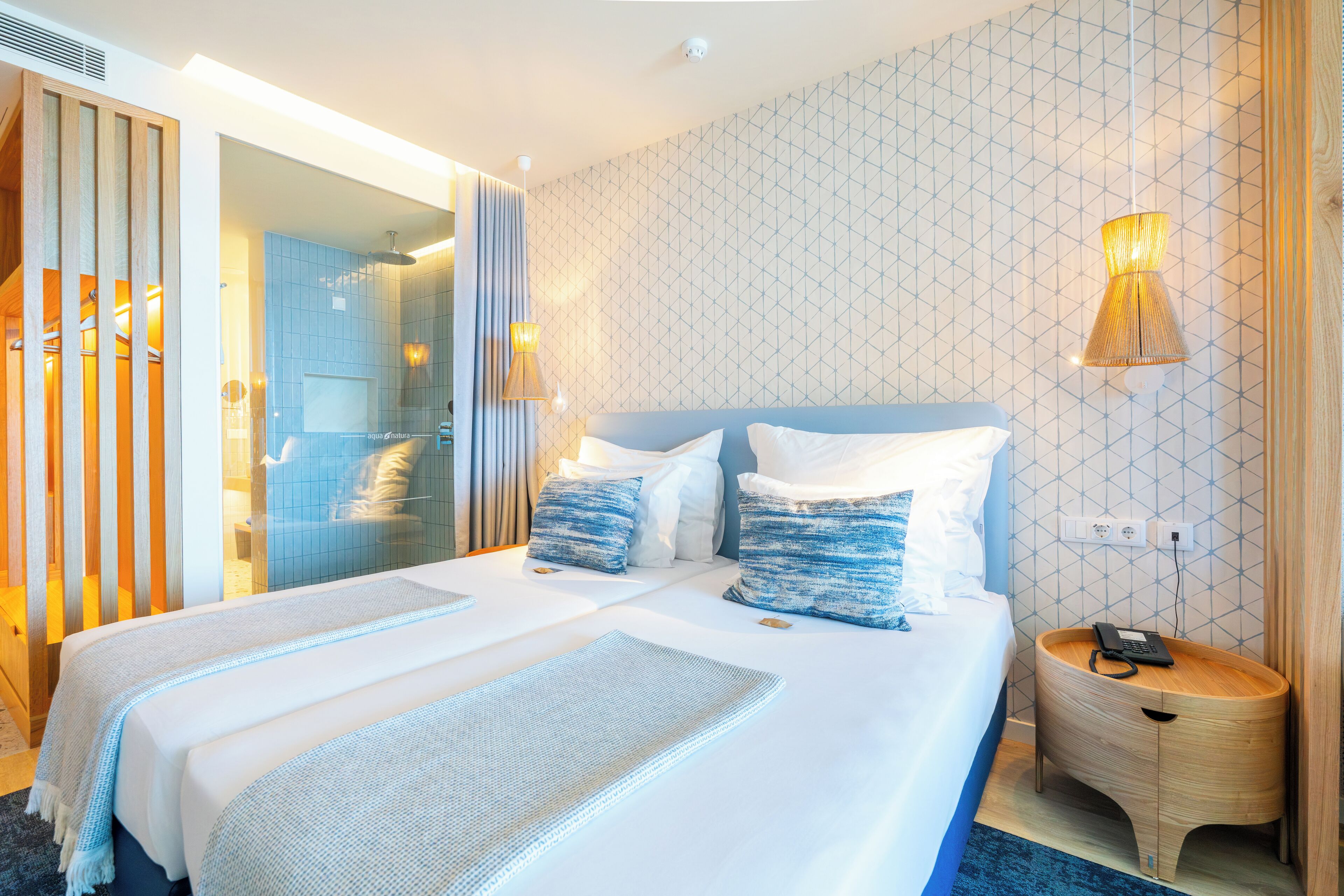 A hotel room with a large bed, a bedside table with a lamp, and a shower area visible through a glass door.