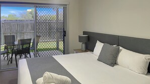 Habitación de hotel con una cama grande, una mesita de noche, una lámpara y vistas a un patio exterior con sillas y una mesa.