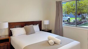 Una cama bien tendida con sábanas blancas y una manta de color beige, un cabecero de madera y una mesita de noche con una lámpara.