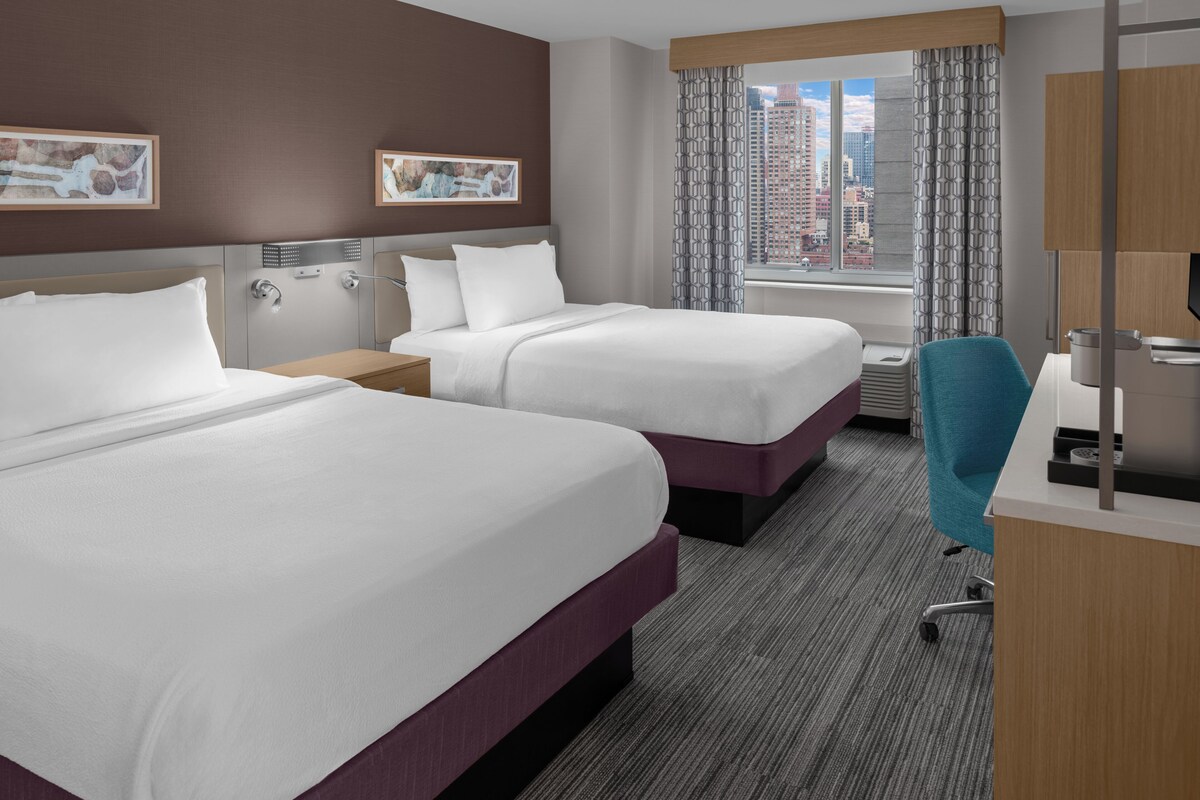 Habitación de hotel con dos camas, un escritorio, una silla, un ventanal con vista a la ciudad y obras de arte en las paredes.