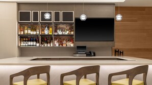 Een bar met drankflessen op planken, een flatscreen televisie en drie houten stoelen met gele kussens.