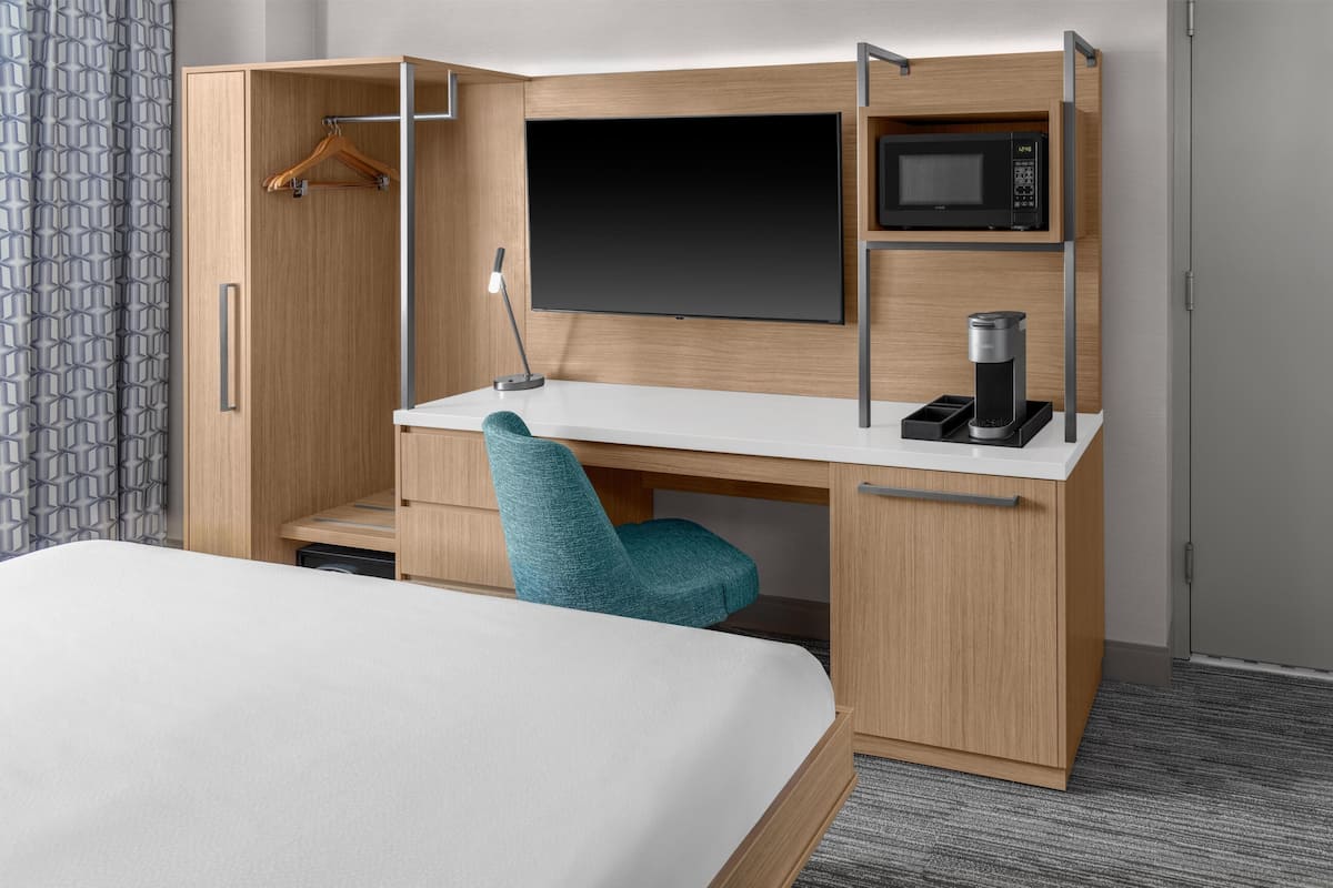 A hotel room with a bed, a desk with a chair, a TV, a microwave, and a coffee machine.