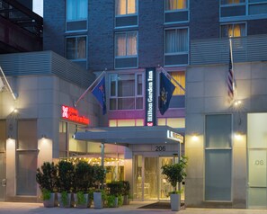 A hotel entrance with a sign reading 'Hilton Garden Inn' and a flag. There are potted plants in front and a covered area with lights. - Hilton Garden Inn New York/Manhattan-Midtown East (New York)